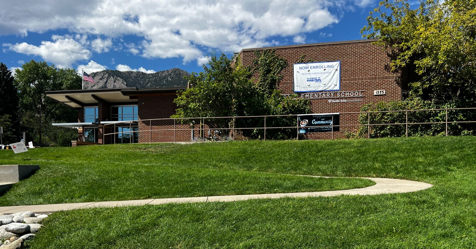 Mesa Elementary’s brick front under a blue sky, with a “Now Enrolling” banner and the Flatirons behind it